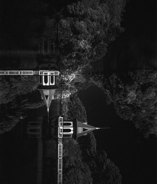 A black and white image shows a building with a pointed roof and a long walkway reflected in water, surrounded by trees.