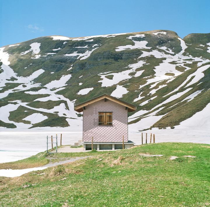 A small pink house with a brown roof sits on a grassy hill next to a frozen lake, with a snow-covered mountain in the background.