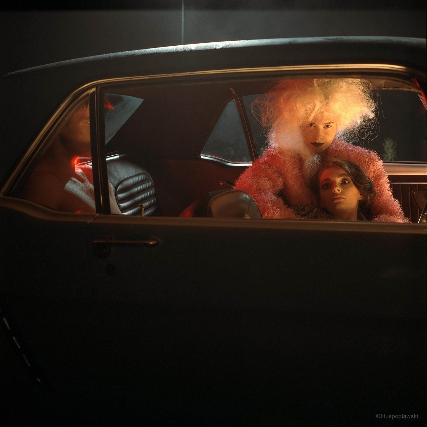 Two women are sitting in a car at night, one with large blonde hair and a pink fluffy coat, the other with dark hair resting on her shoulder.