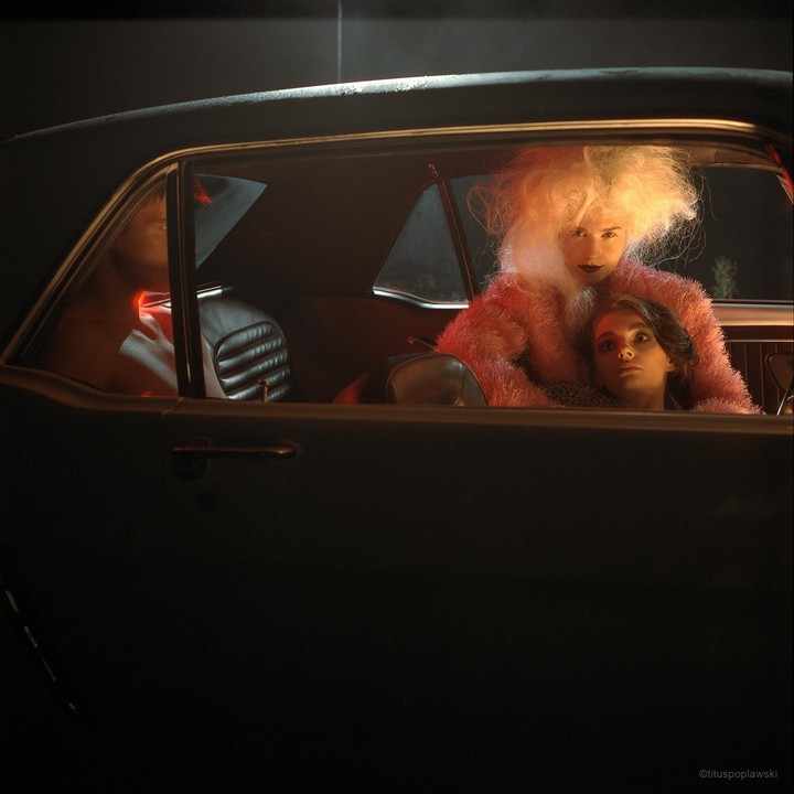Two women are sitting in a car at night, one with large blonde hair and a pink fluffy coat, the other with dark hair resting on her shoulder.
