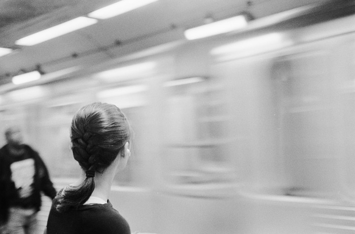 A woman with a braided ponytail stands on a subway platform as a train speeds past.