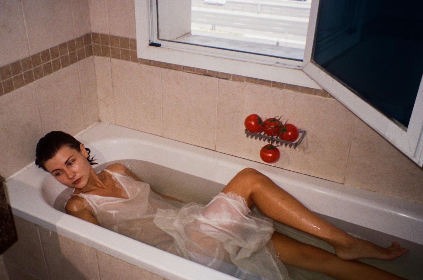 A woman in a white slip lies in a bathtub with tomatoes on a soap dish nearby.