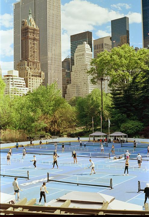 People play pickleball on blue courts surrounded by trees and tall buildings.