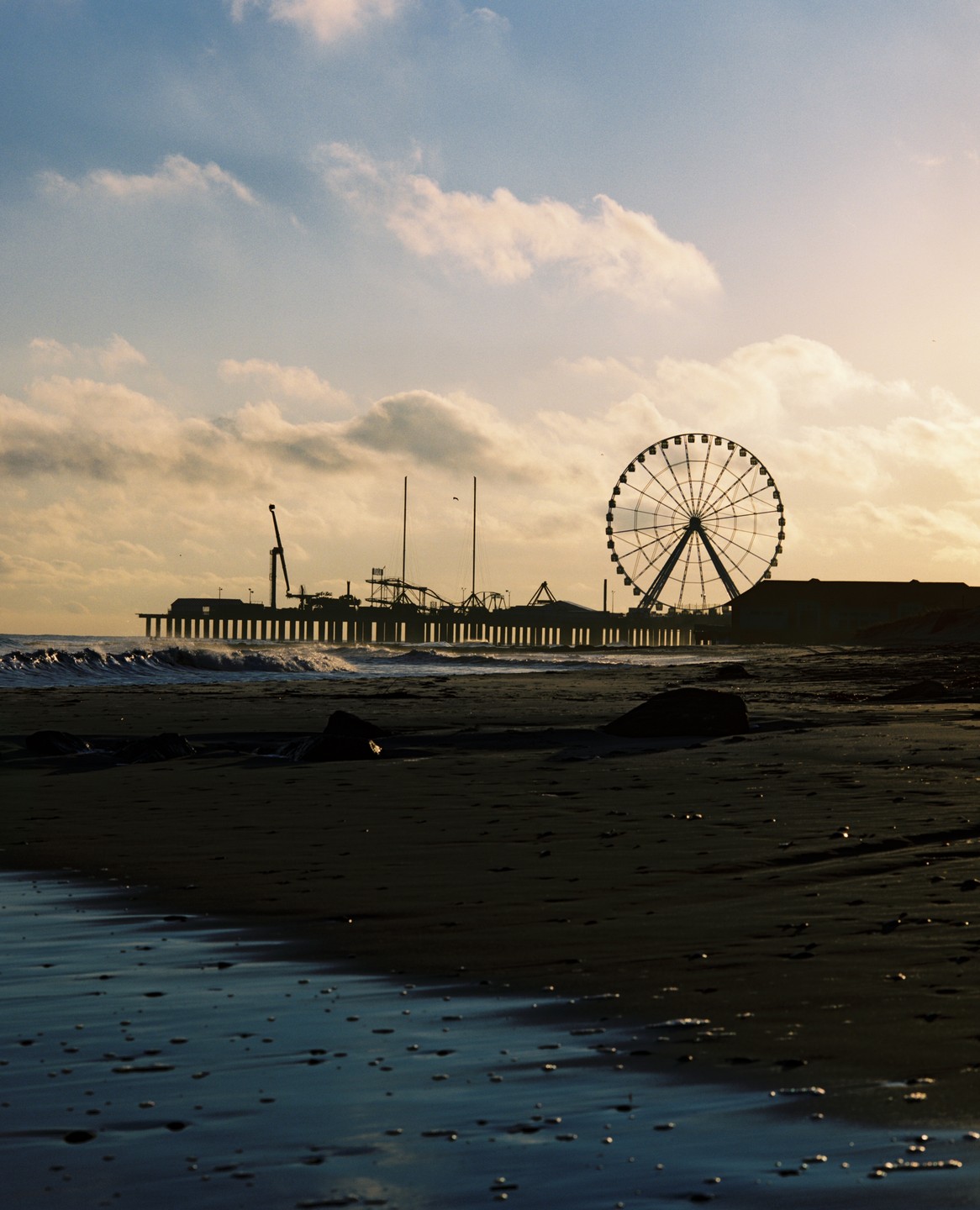 A ferris wheel and pier are silhouetted against a cloudy sky at dusk, with waves breaking on the beach in the foreground.