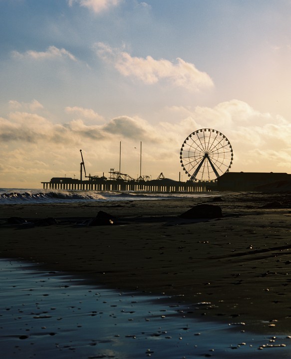 A ferris wheel and pier are silhouetted against a cloudy sky at dusk, with waves breaking on the beach in the foreground.