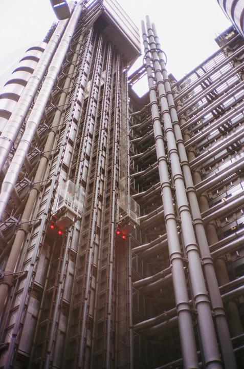 Two glass elevators with red lights are visible on the side of a modern building with many pipes and tubes.
