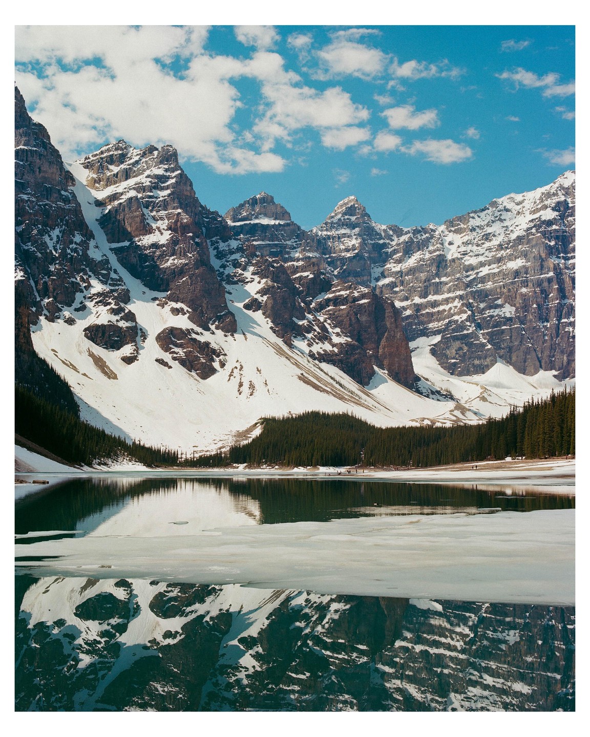 Snow-capped mountains reflect in a partially frozen lake surrounded by pine trees under a blue sky with clouds.