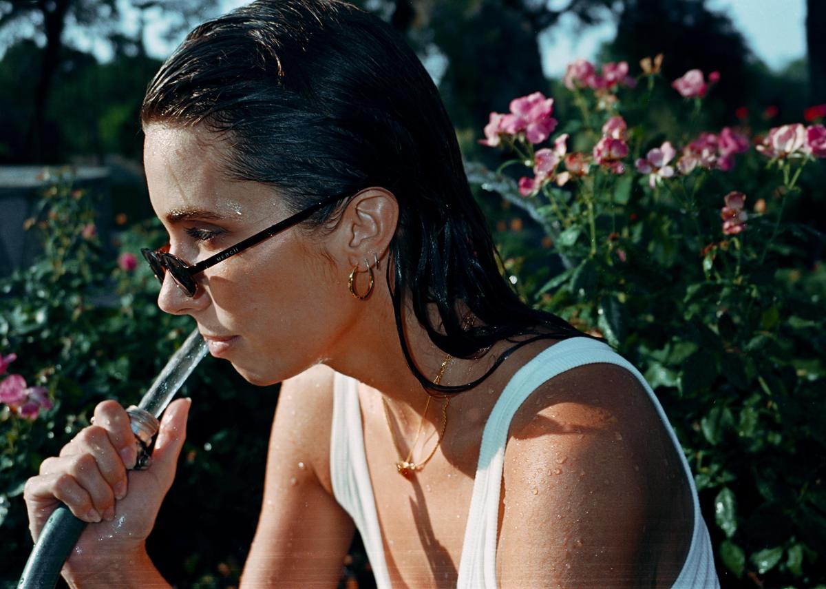 A woman in sunglasses drinks from a garden hose in a rose garden.