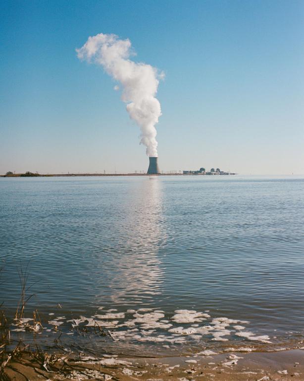 A power plant with a tall smoke stack emitting a large plume of smoke stands across a body of water under a clear sky.