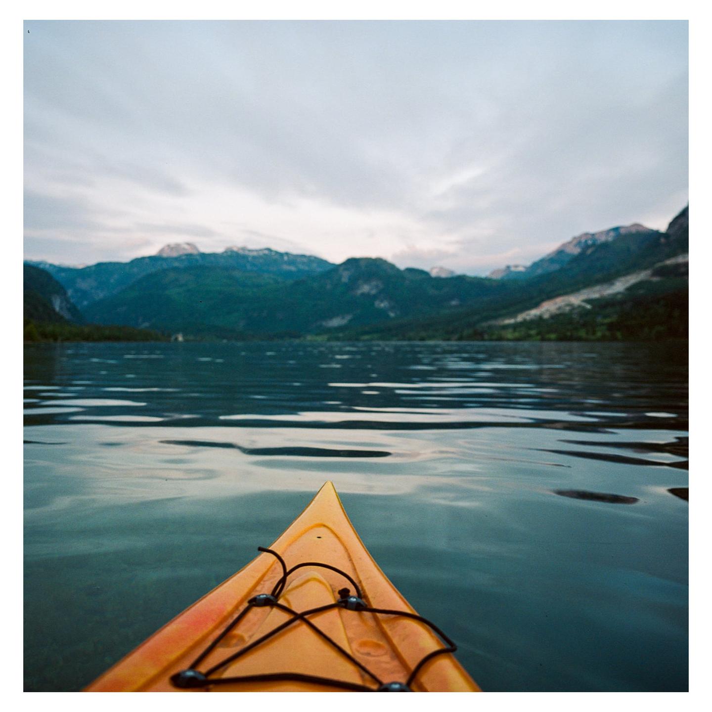 The bow of a yellow kayak points towards a lake surrounded by mountains under a cloudy sky.