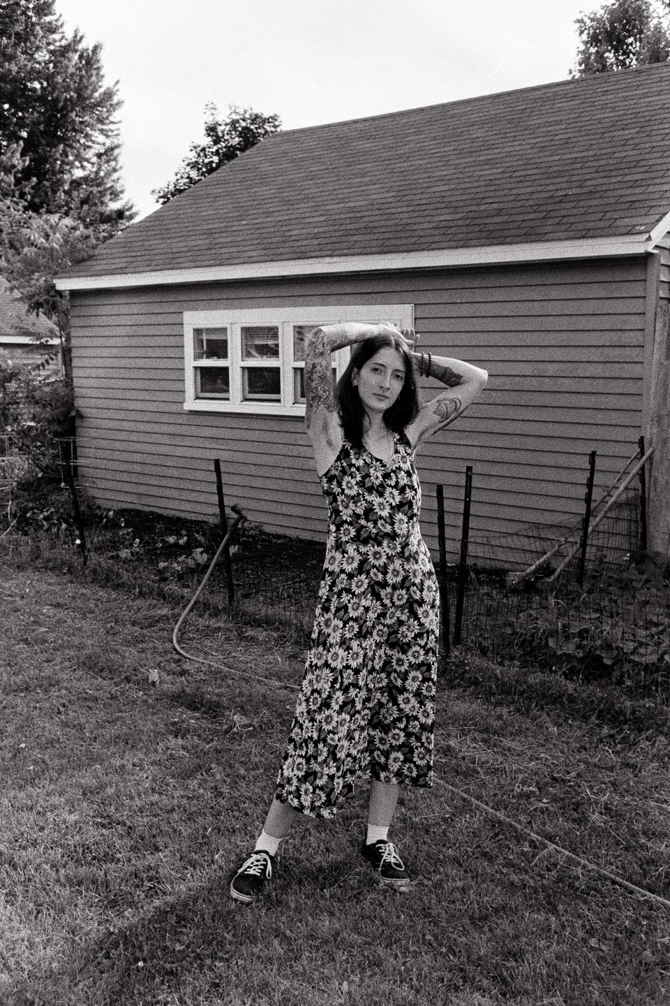 A woman with tattooed arms stands in a grassy yard in front of a house.