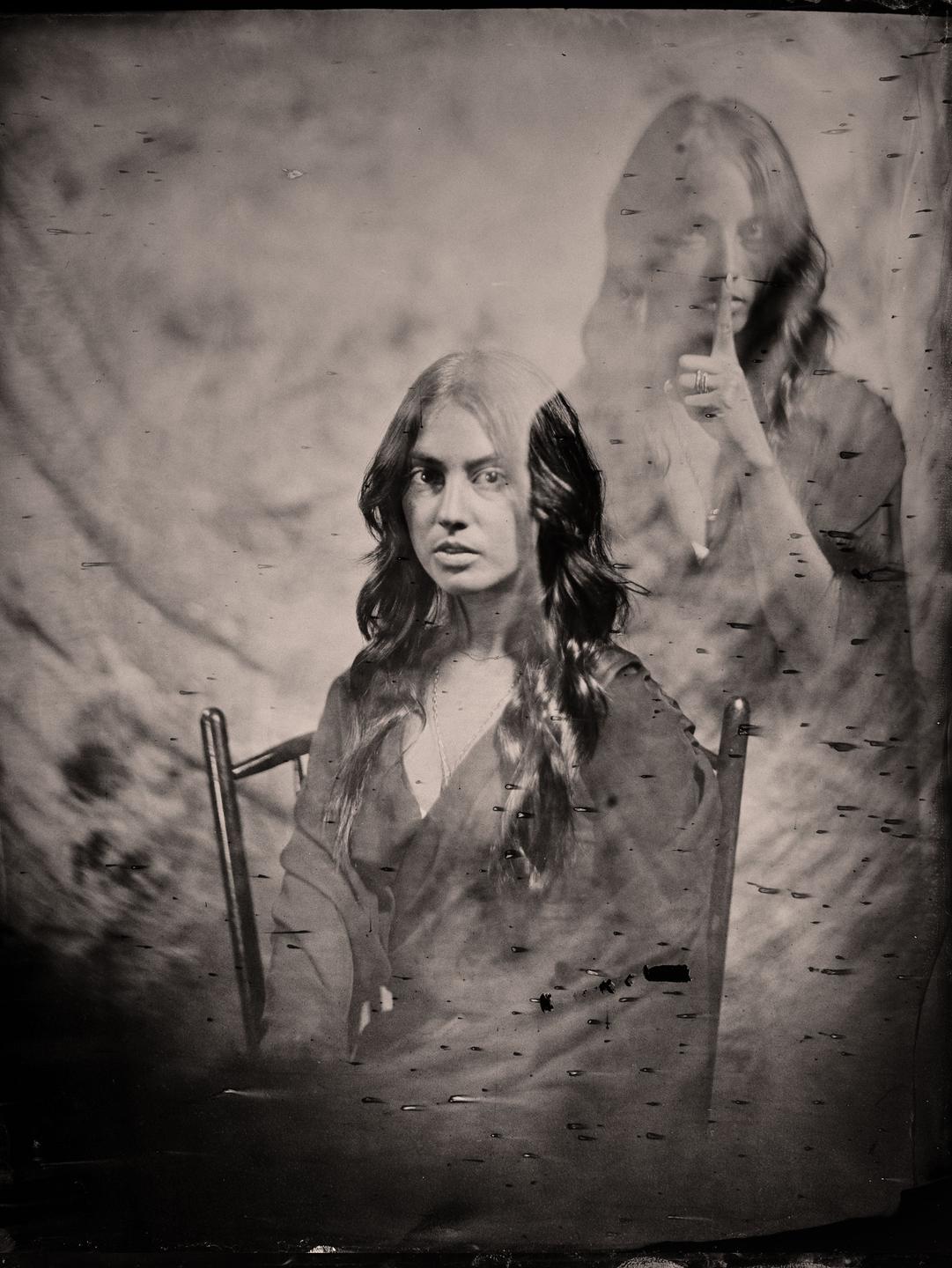 A woman with long hair sits in a chair, with a ghostly double exposure of herself in the background holding a finger to her lips.