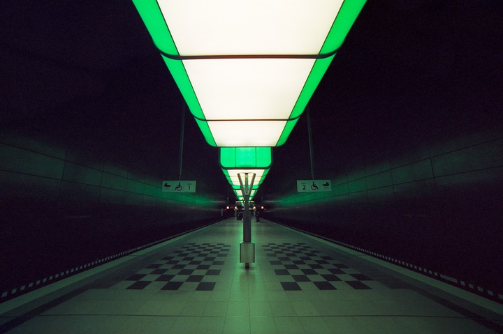 A subway platform is illuminated by green and white lights on the ceiling.