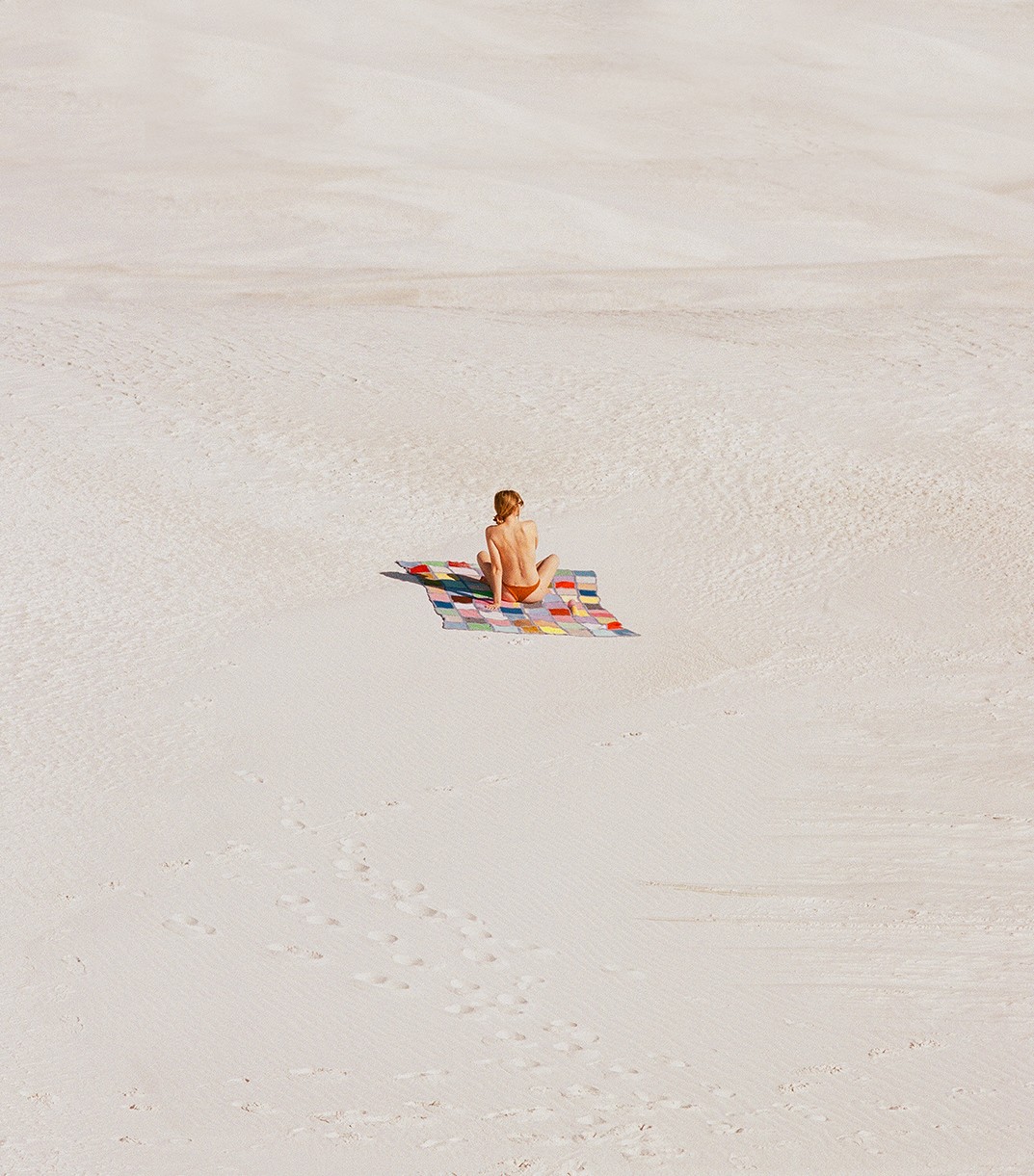 A woman sits on a colorful blanket in the middle of a vast expanse of sand.