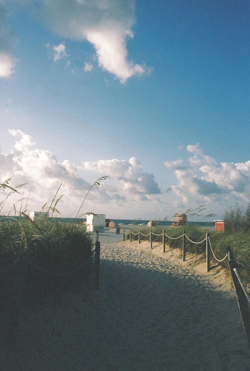 A sandy path leads to the ocean, lined with rope fences and tall grass under a blue sky with clouds.