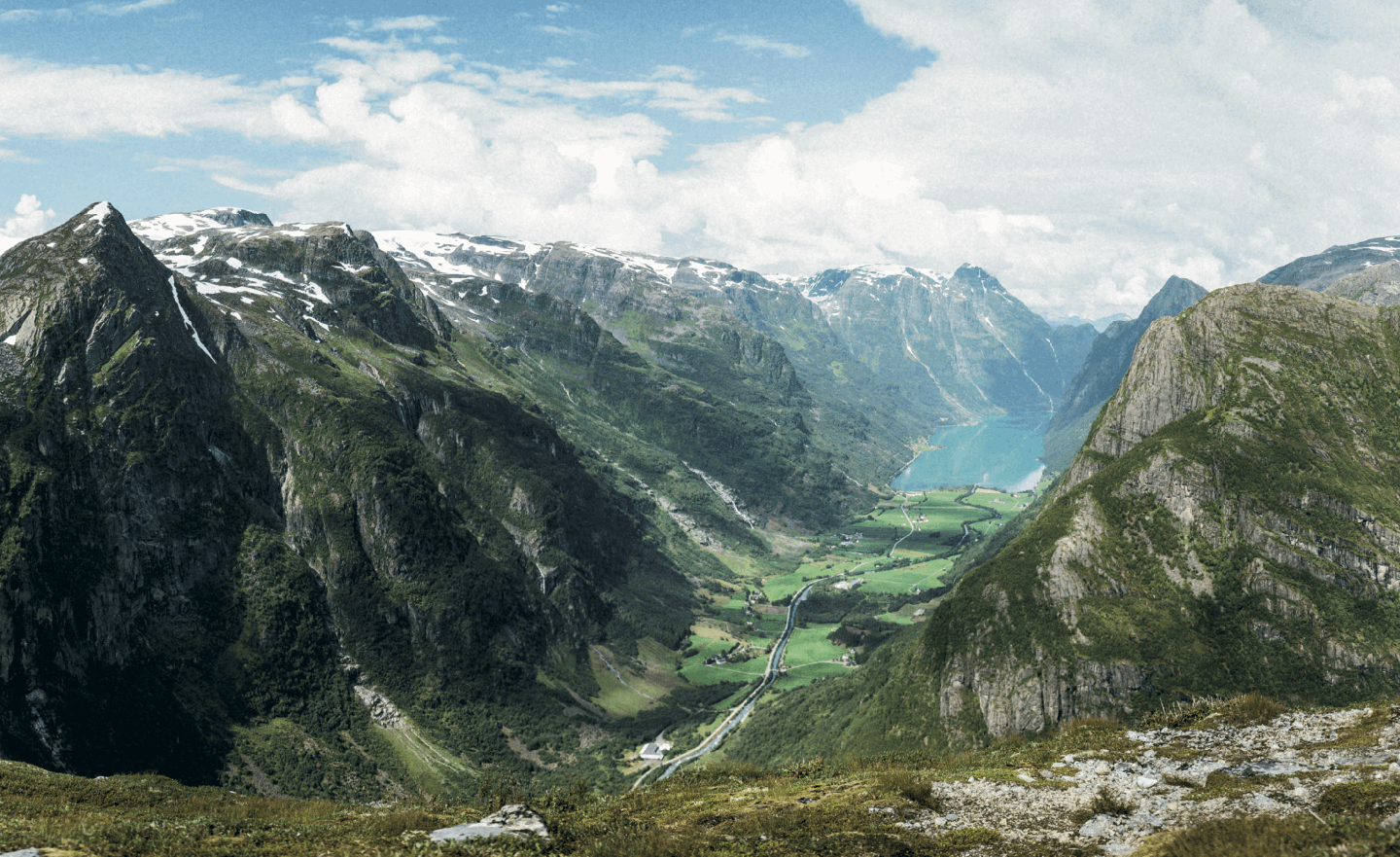 A vast mountain landscape features a valley with a river and a lake, surrounded by snow-capped peaks under a cloudy sky.