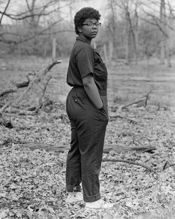 A woman wearing glasses and a jumpsuit stands in a forest with fallen leaves on the ground.