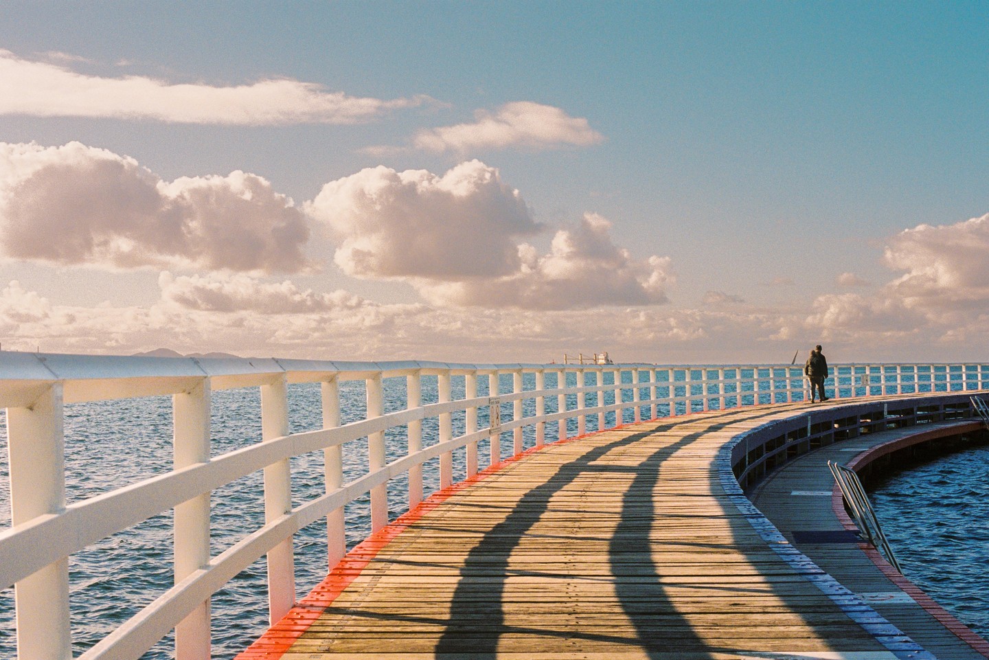 A person walks along a curved wooden pier with white railings over blue water under a partly cloudy sky.