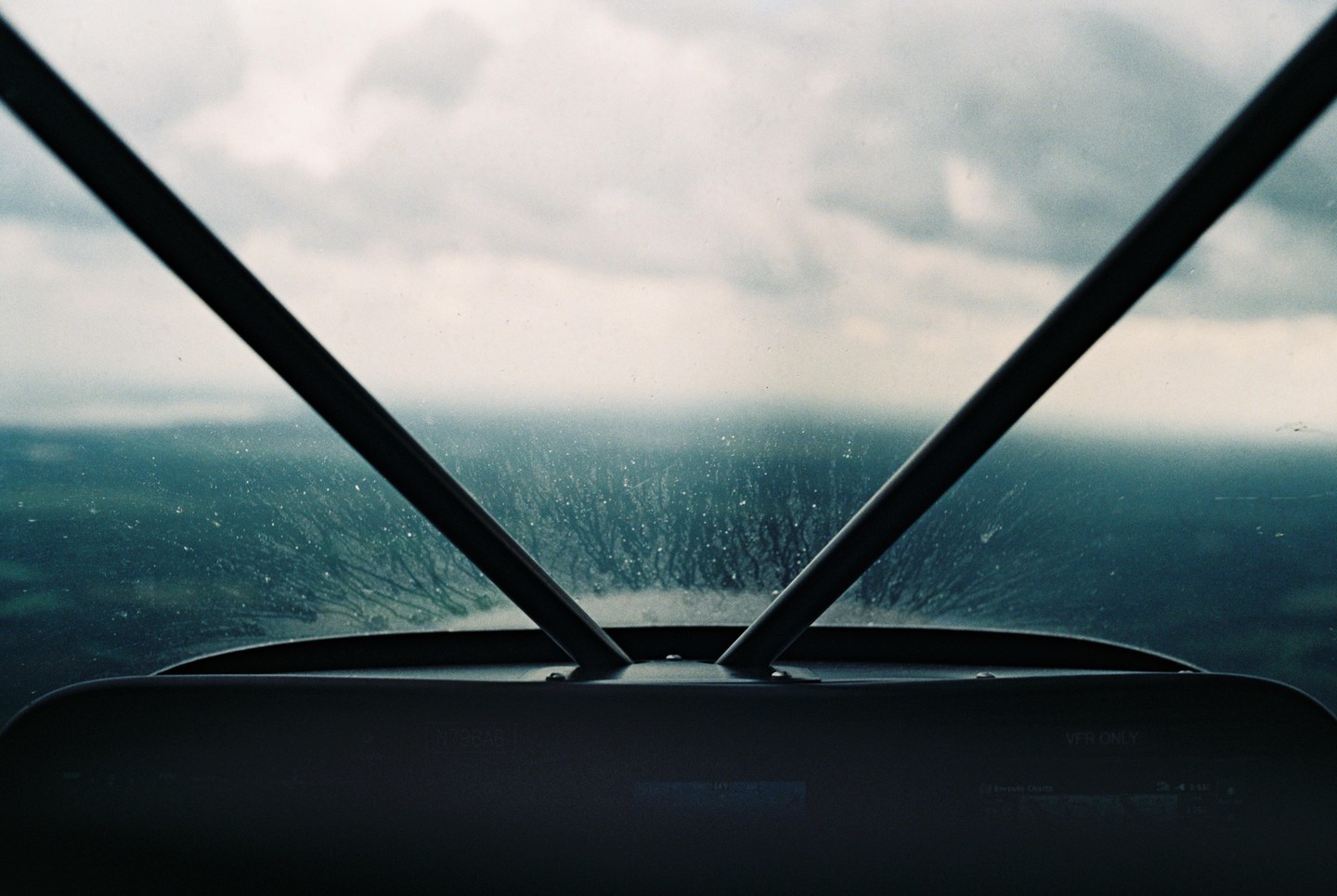 The view from a cockpit shows a cloudy sky and a landscape with trees through a rain-splattered windshield.