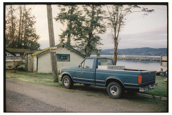 A blue pickup truck is parked on a gravel road next to a small building with a body of water and mountains in the background.