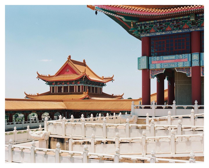 Two ornate temple buildings with red roofs and colorful details are visible behind white stone railings under a clear sky.