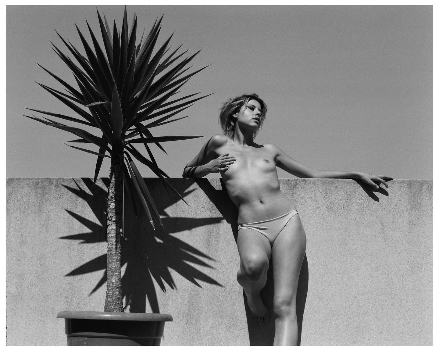 A woman in underwear leans against a wall next to a potted yucca plant.