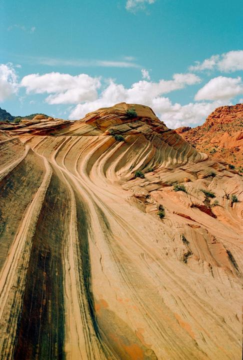 A desert landscape shows a large rock formation with wavy, striped patterns and a blue sky with white clouds.