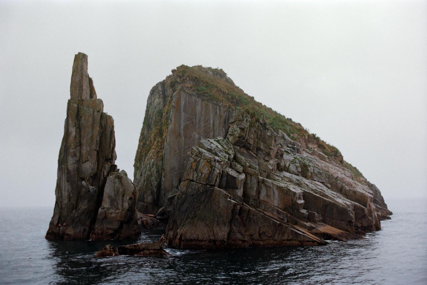 Two large rock formations rise from the ocean under a hazy sky.