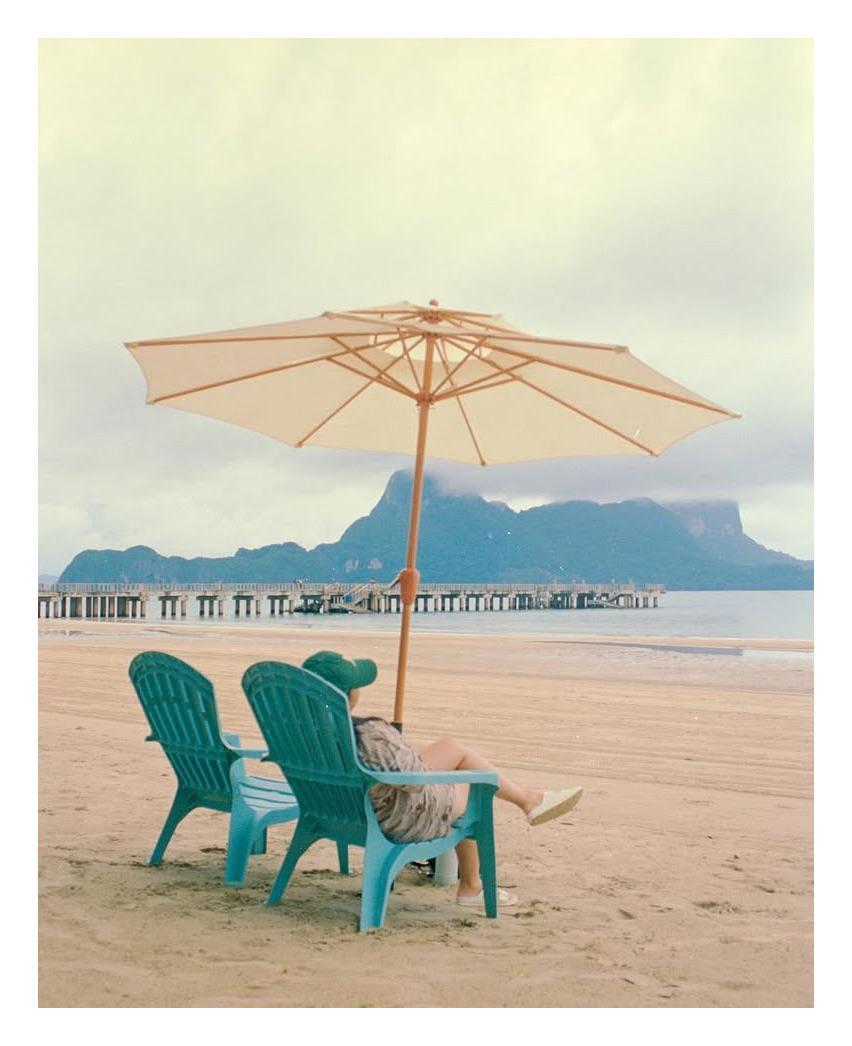 A woman sits on a beach chair under an umbrella, looking out at the ocean and mountains.