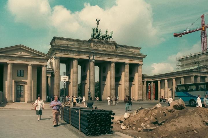The Brandenburg Gate stands tall with a construction crane visible in the background and people walking in the foreground.