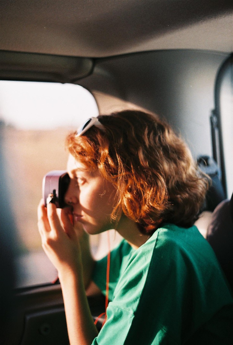 A woman with orange hair and sunglasses on her head looks out a car window while holding a camera.
