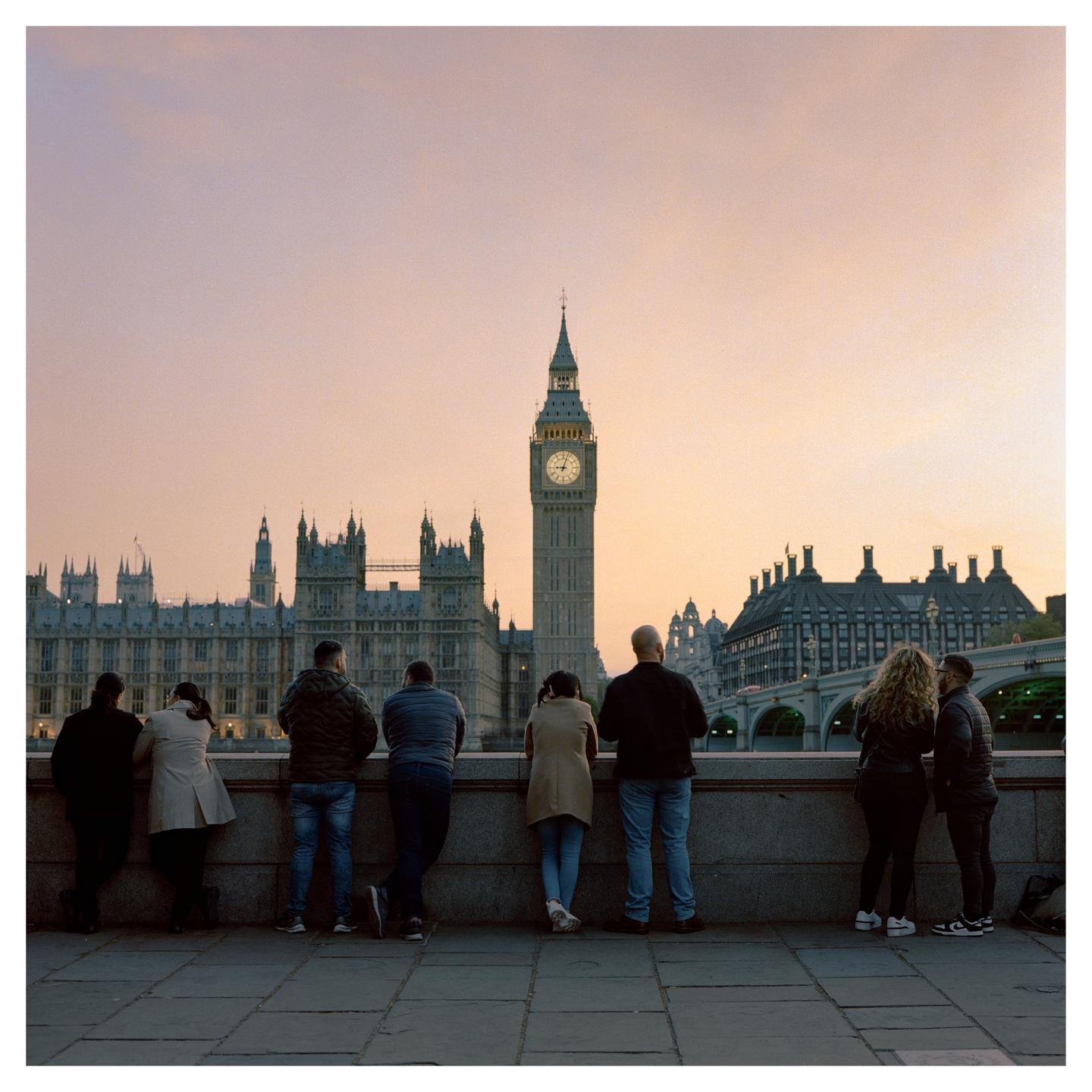 Six people stand with their backs to the viewer, looking at the Houses of Parliament and Big Ben at sunset.