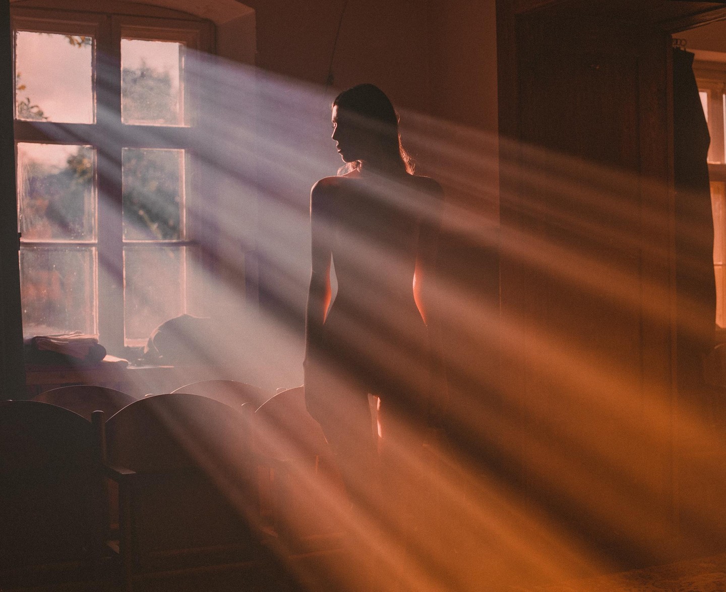 A woman stands in a room with sunbeams shining through a window.