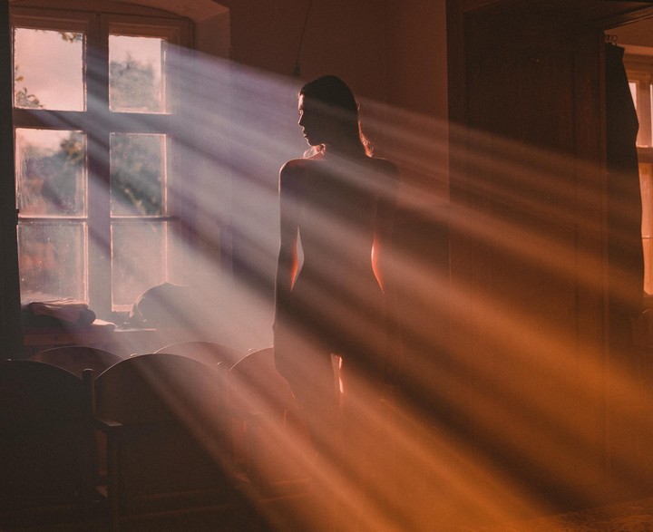 A woman stands in a room with sunbeams shining through a window.