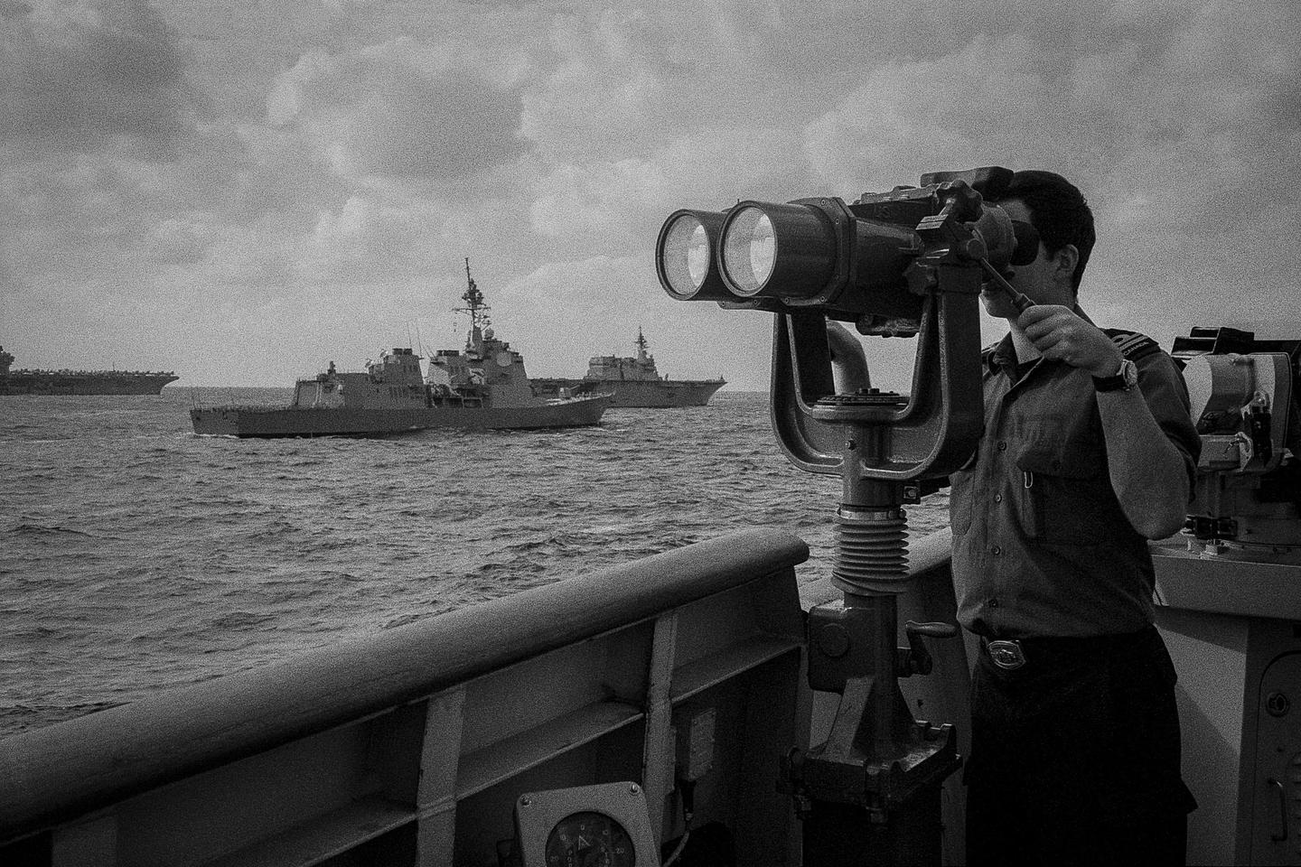 A man looks through large binoculars on a ship at sea, with other ships visible in the distance under a cloudy sky.