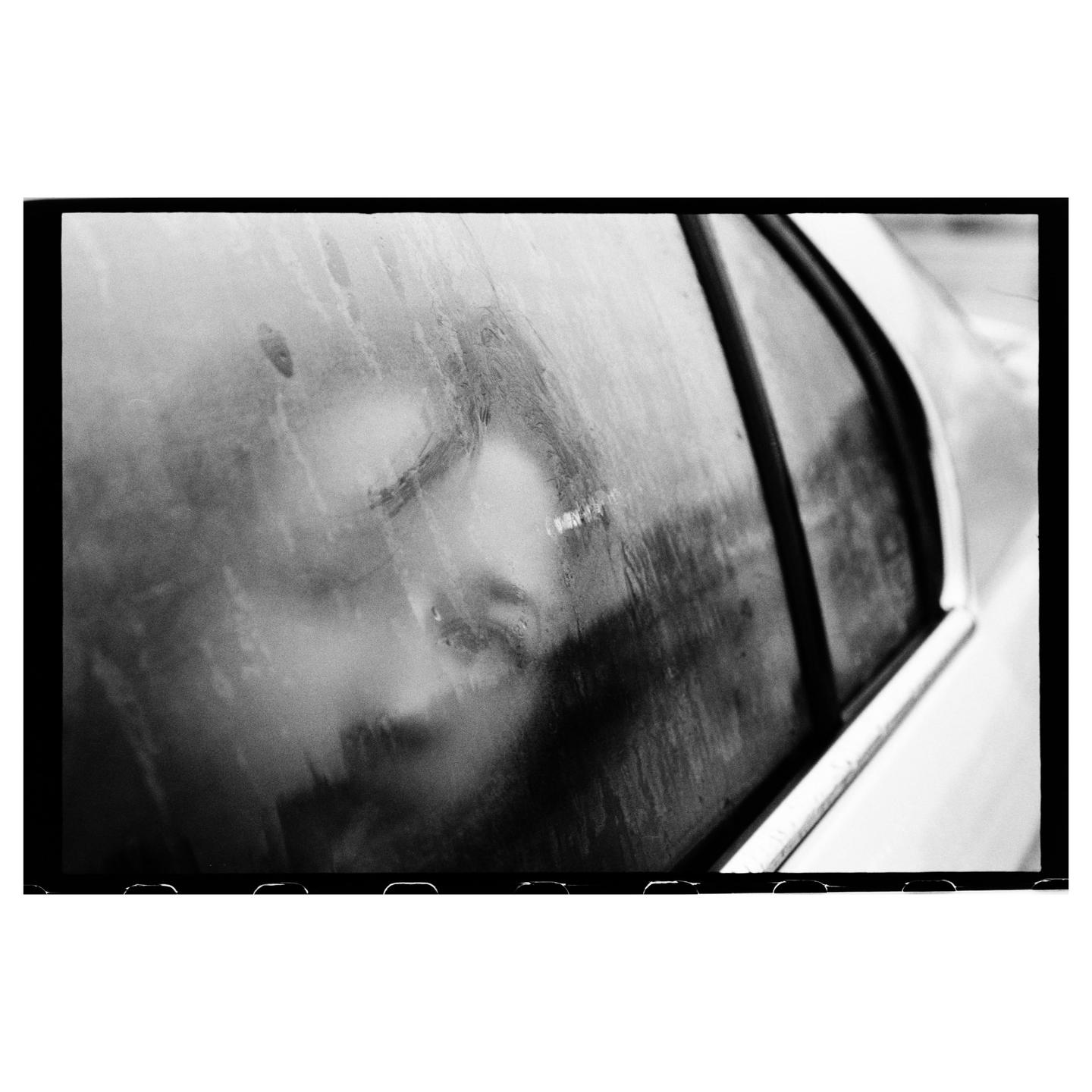 A person's face is visible through a wet car window.