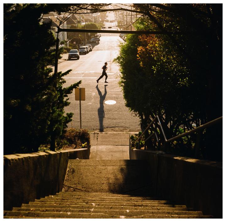 A man runs across a sunlit street at the bottom of a long concrete staircase.