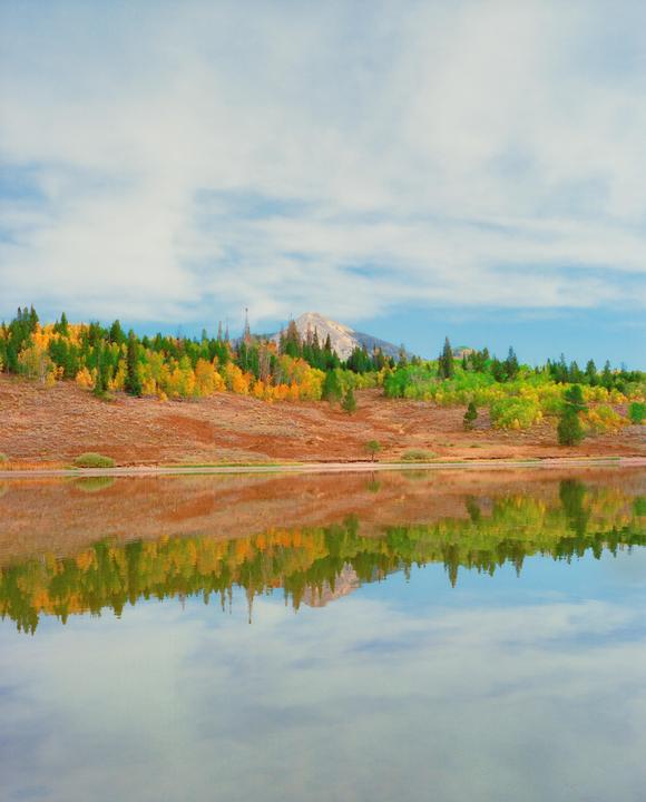 A mountain with autumn foliage is reflected in a still lake under a cloudy sky.