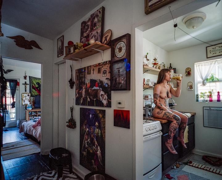 A nude woman with tattoos sits on a kitchen counter and drinks from a jar.