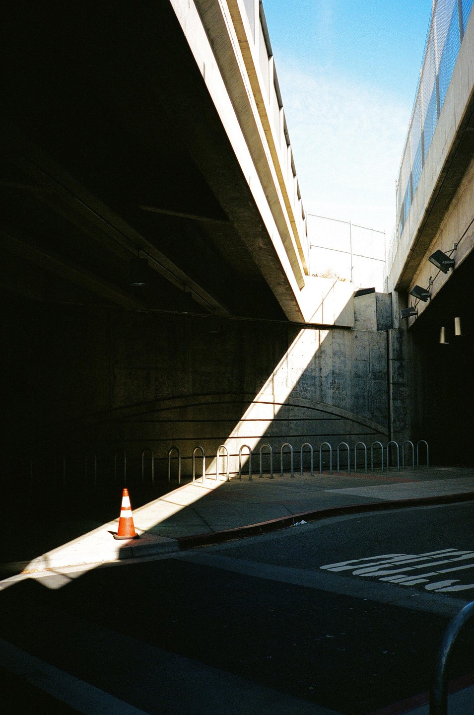 A traffic cone sits in a beam of sunlight under an overpass next to a row of bicycle racks.