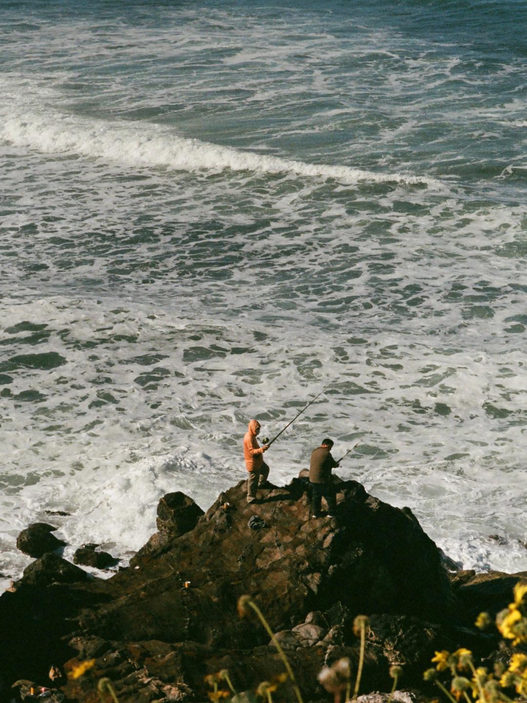 Two men are fishing from a rocky outcrop as waves crash around them.