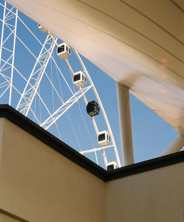 A large white ferris wheel with several gondolas is visible against a clear blue sky, framed by architectural elements.