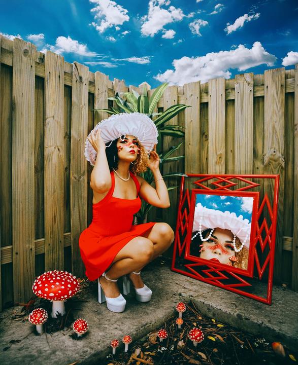 A woman in a red dress and white hat crouches next to a mirror and red and white mushrooms.