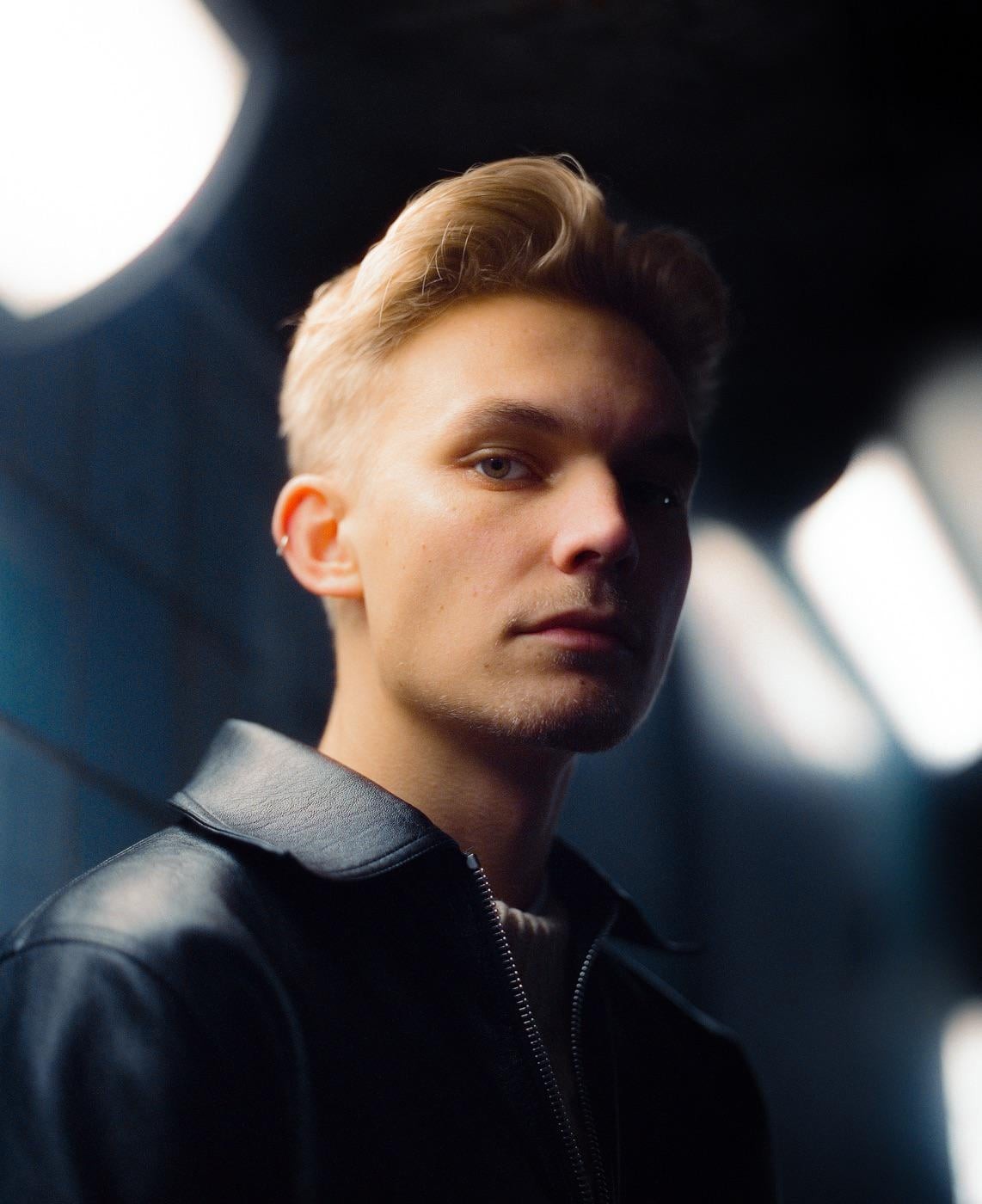 A man with blond hair and a black leather jacket looks directly at the viewer.