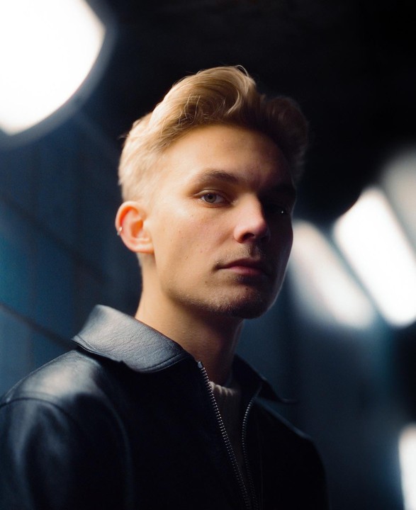 A man with blond hair and a black leather jacket looks directly at the viewer.