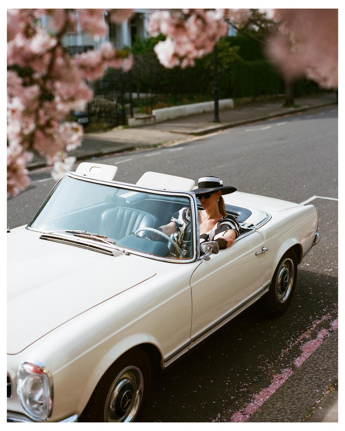 A woman in a wide-brimmed hat and sunglasses drives a white convertible on a street lined with cherry blossoms.