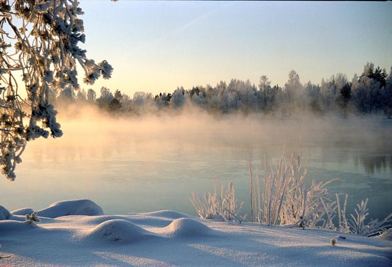 Mist rises from a river with snow-covered banks and trees under a clear sky.