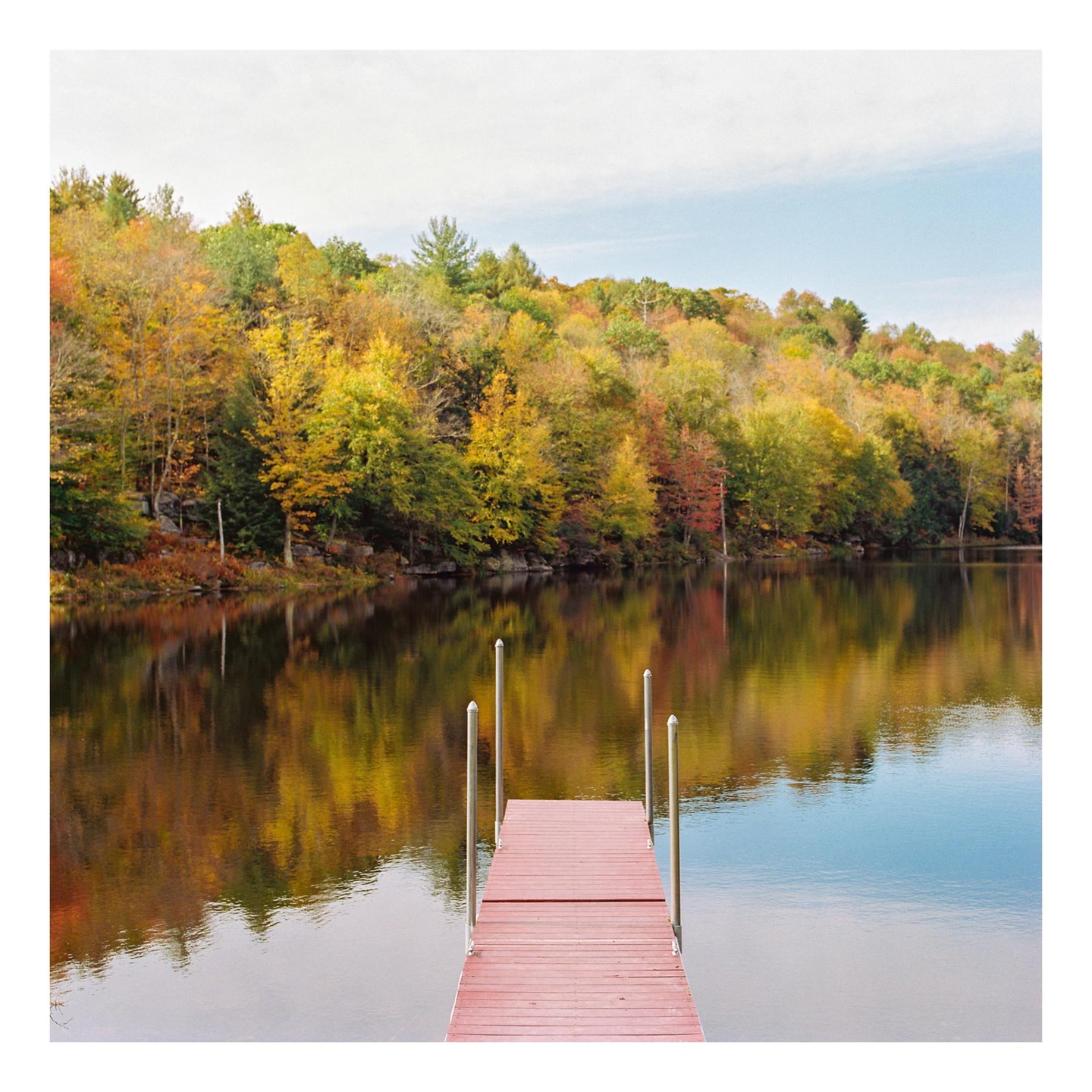 A red dock extends into a calm lake reflecting autumn trees under a bright sky.