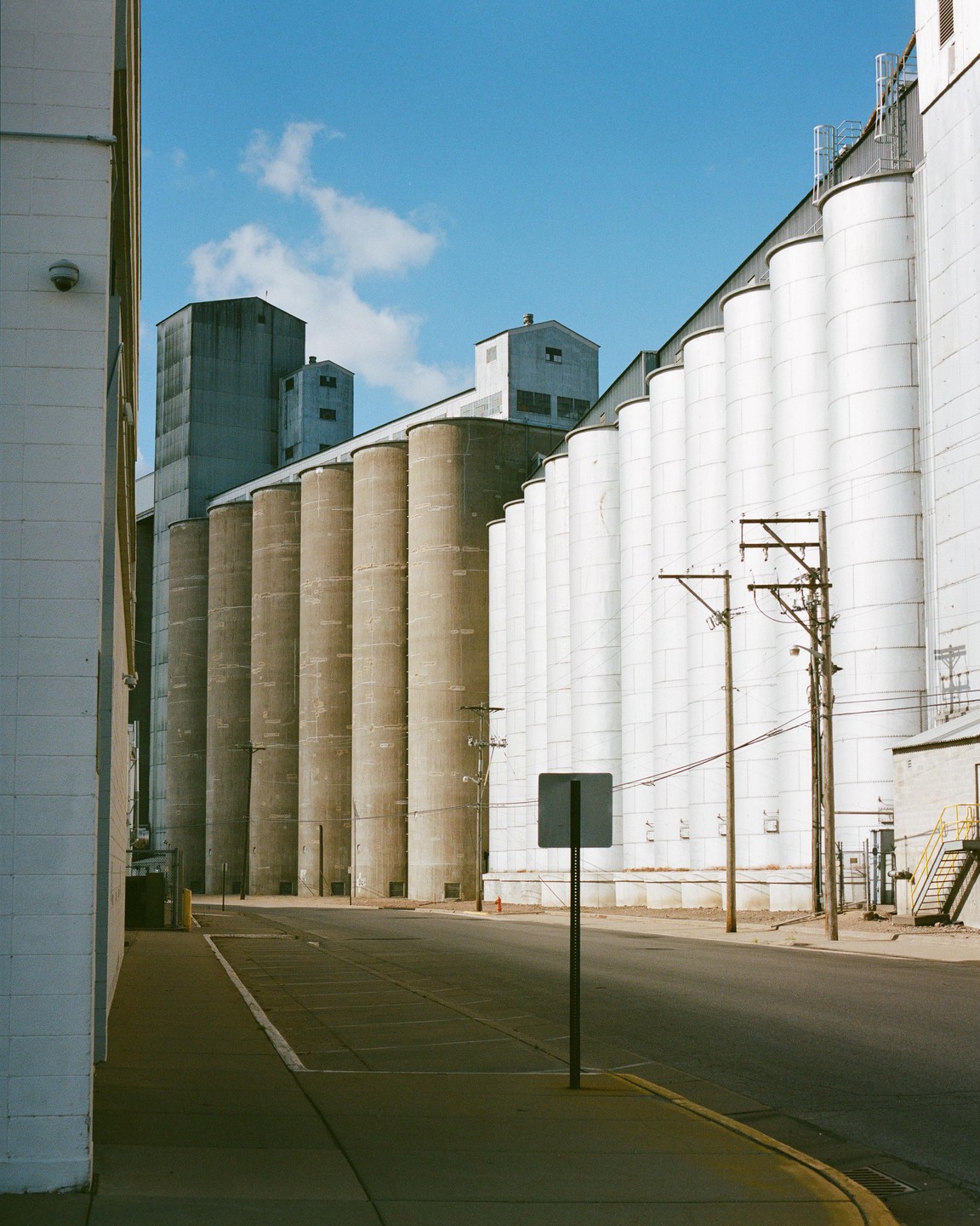 A street runs between a building and a row of grain silos under a blue sky.