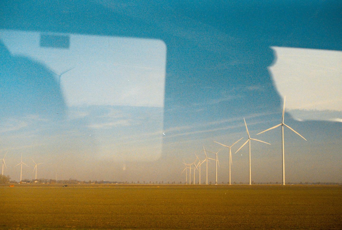A row of wind turbines stands in a field under a blue sky, with reflections visible in the upper part of the image.